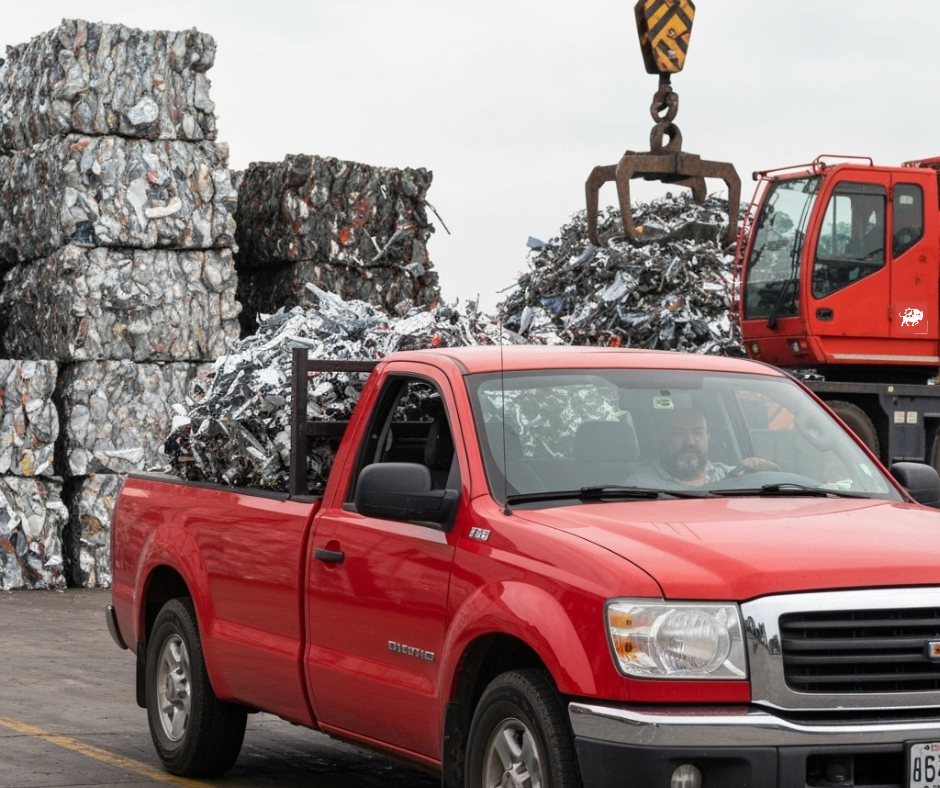 Red pickup truck delivering scrap metal to a recycling yard with stacked metal bales and a crane in the background.